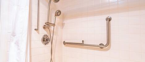 A bathroom with white tiled walls, a shower with a handheld showerhead, a handheld showerhead, and a grab bar.