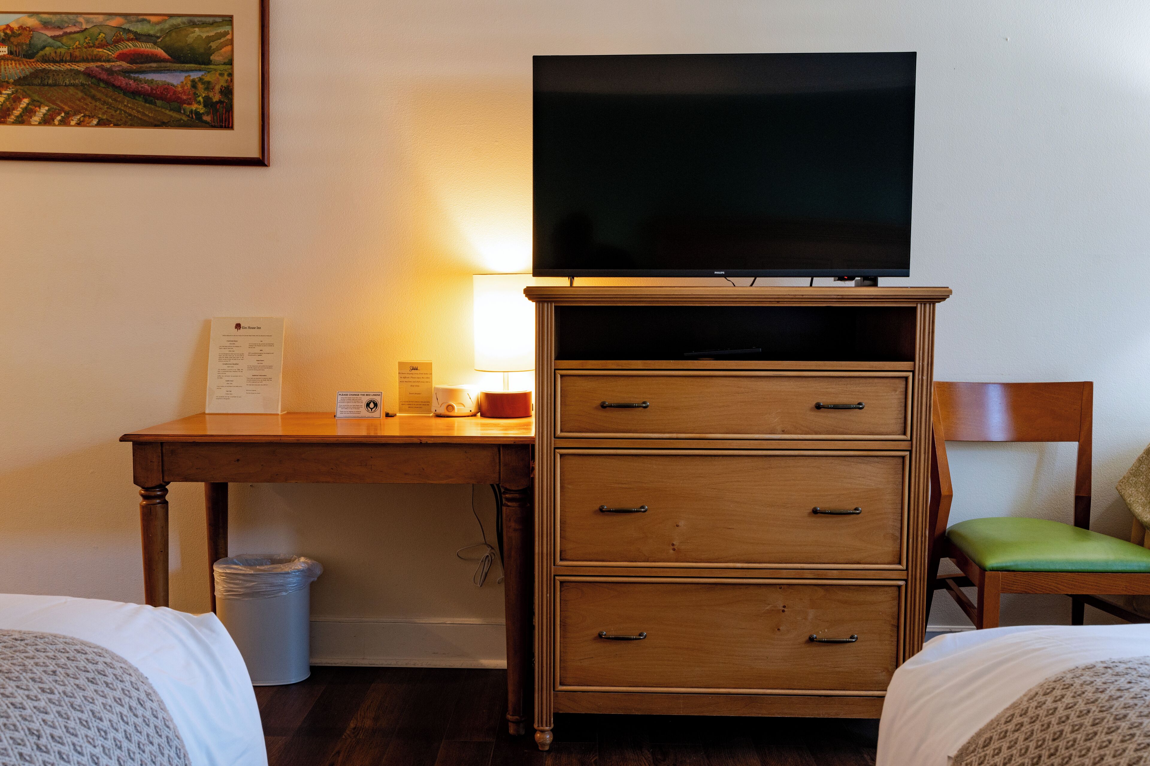 Habitación de hotel con cómoda de madera, televisor de pantalla plana encima, escritorio con lámpara, una silla y una cama.