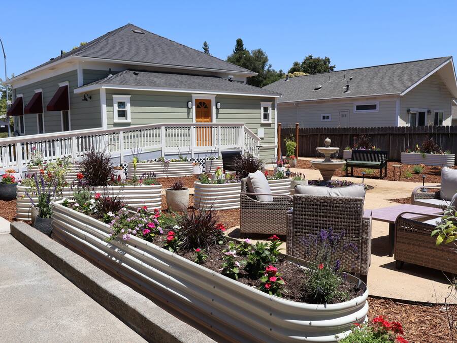 A residential area with a house featuring a porch with outdoor seating, a garden with various plants and flowers, and a wooden fence.