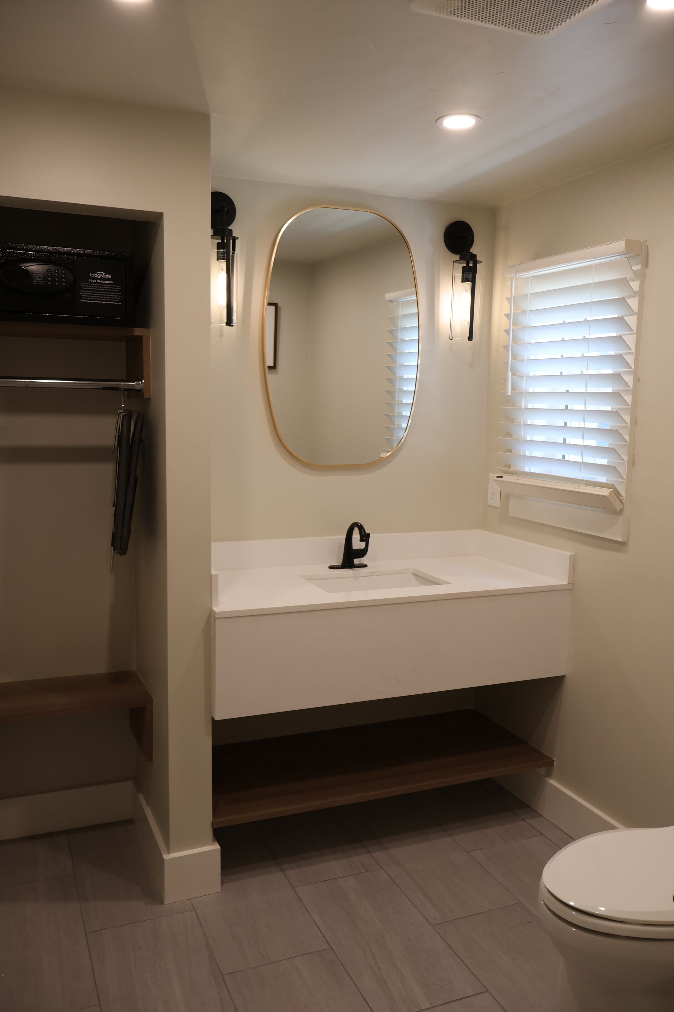 A bathroom with a white sink, a mirror, and a toilet. There is a shelf above the sink and a window with blinds.
