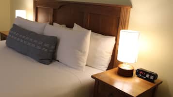 Una cama bien tendida con almohadas blancas y grises, un cabecero de madera, una lámpara de noche y un reloj digital sobre una mesita de noche de madera.
