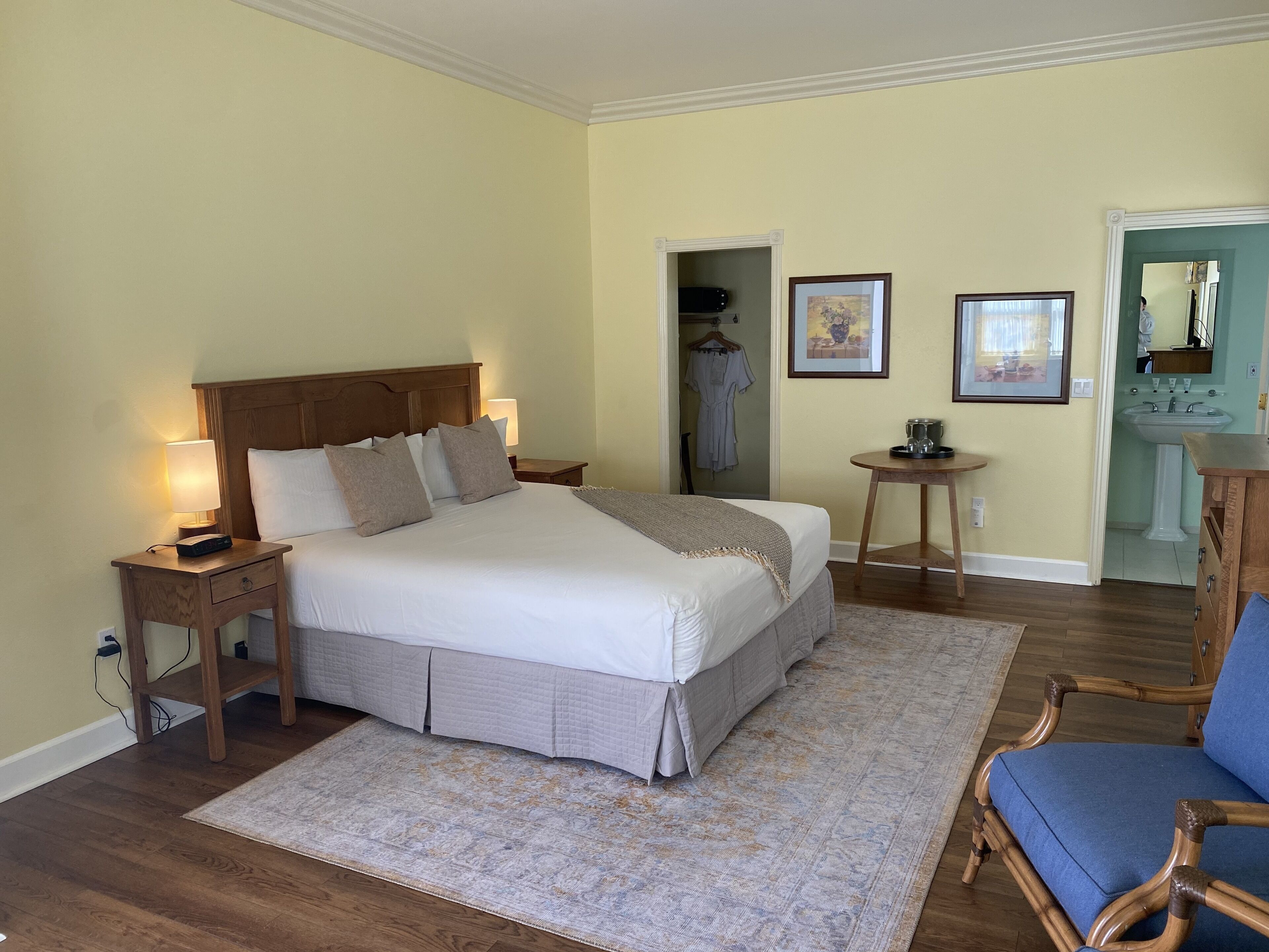Una habitación de hotel con una cama grande, mesitas de noche, una pequeña mesa redonda, una silla, un baño con lavamanos y cuadros en la pared.