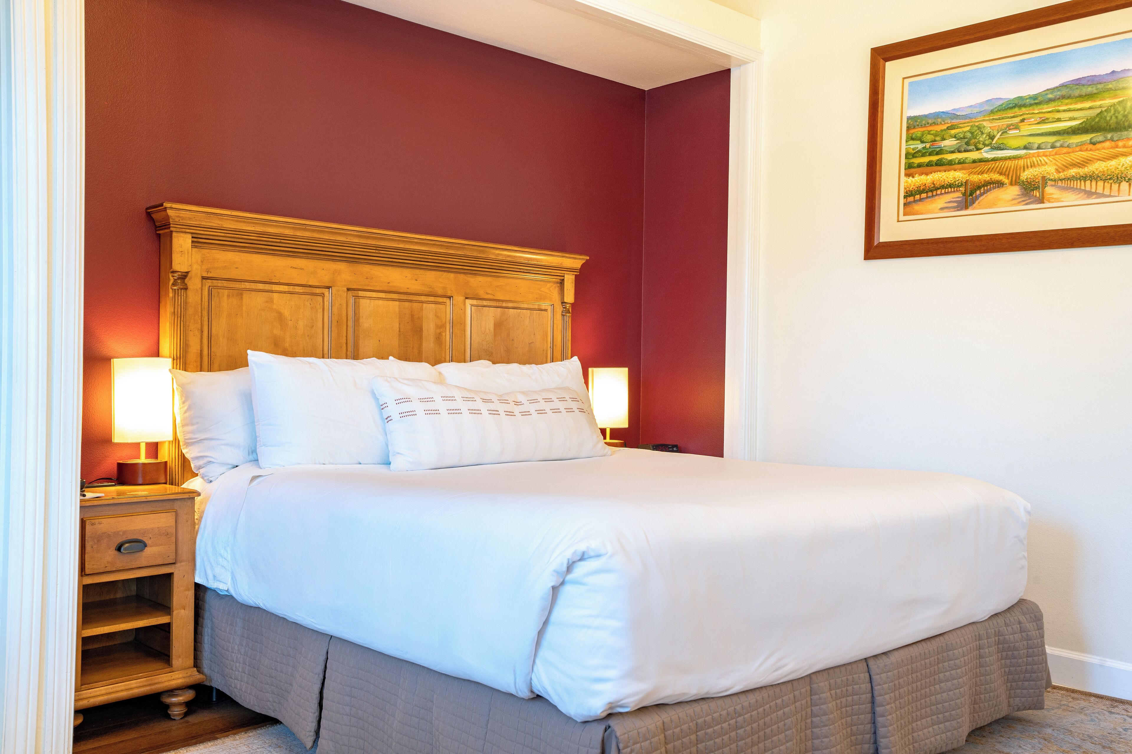 A neatly made bed with white linens and a wooden headboard, a bedside table with a lamp, and a framed landscape painting on the wall.