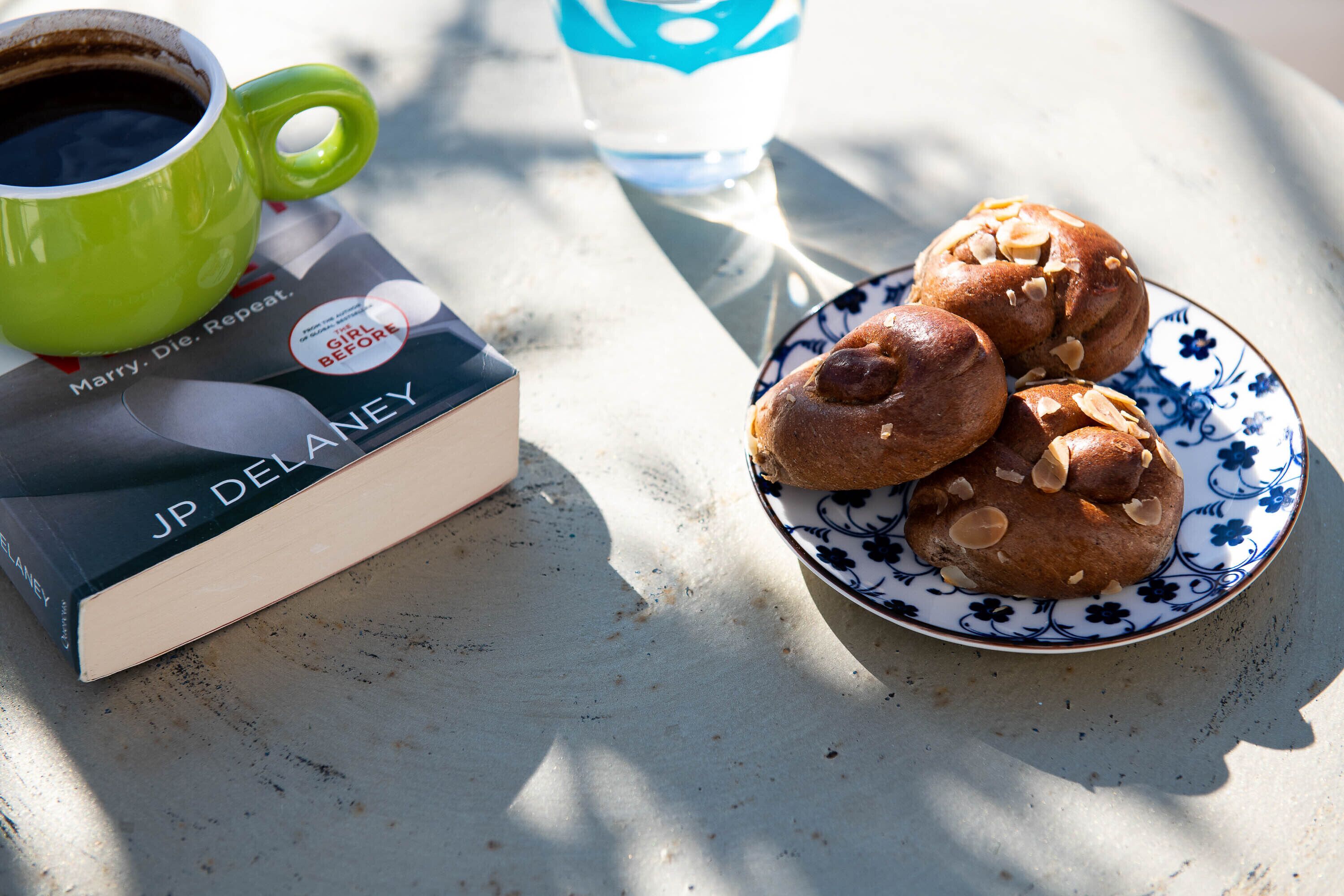 A book titled "Girl Before" by JP Delaney, a green mug, a plate with pastries, and a water bottle on a table.