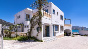 Um edifício branco com varandas e uma porta azul, cercado por um espaço pavimentado e vegetação.