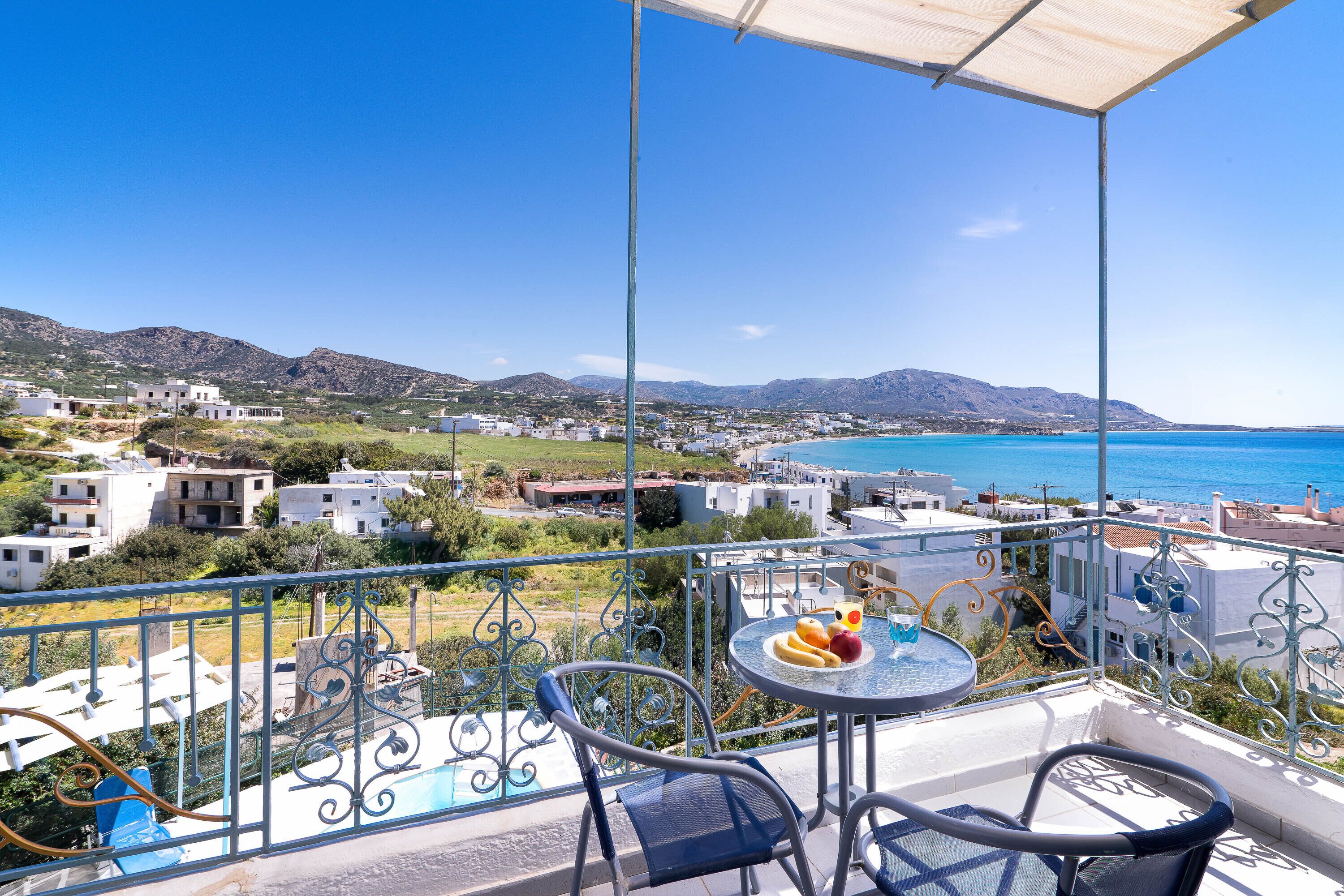 A balcony with a table and chairs, overlooking a coastal town and the sea.