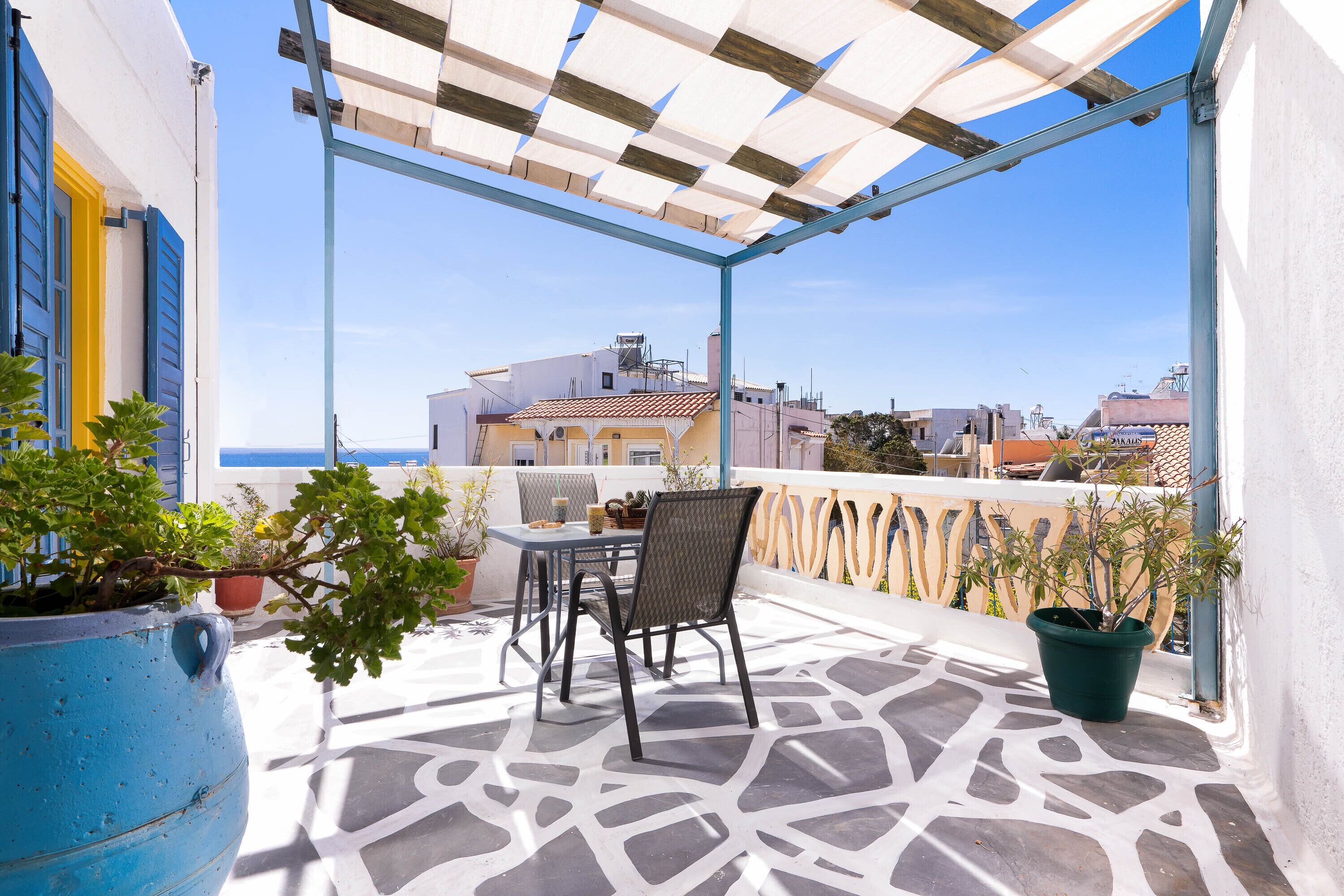 Terraço com mesa e cadeiras, plantas em vasos e vista para o mar e edifícios.