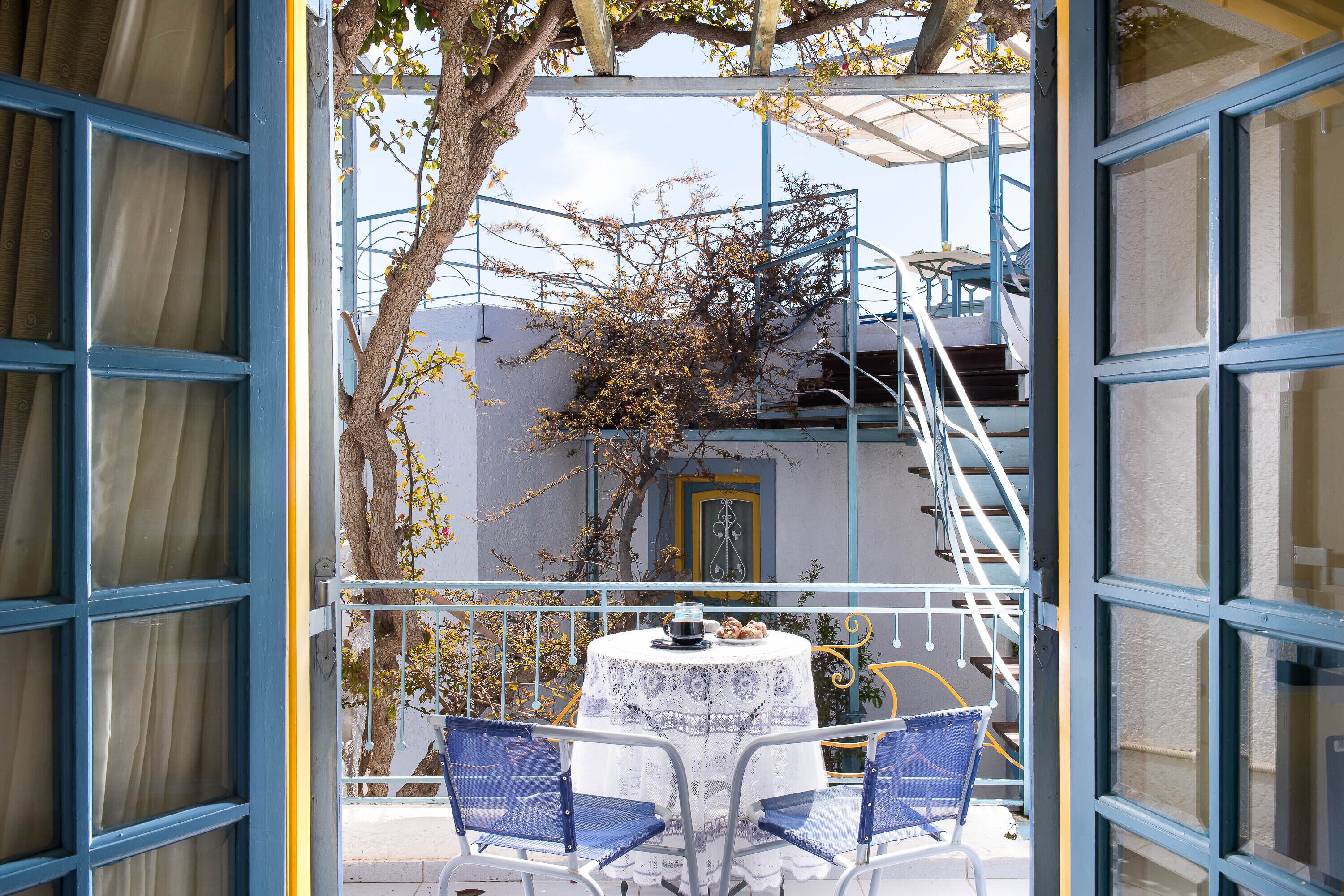 Un balcon ouvert avec une table et des chaises, une vue sur un immeuble avec une porte jaune, et un arbre aux branches nues.