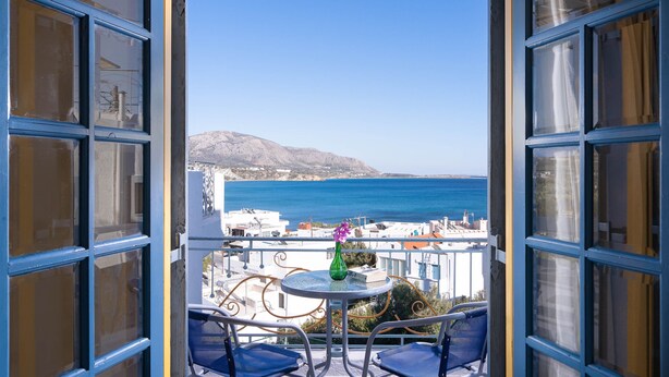 Varanda aberta com vista para o mar e uma montanha, com uma mesa, um vaso e duas cadeiras.