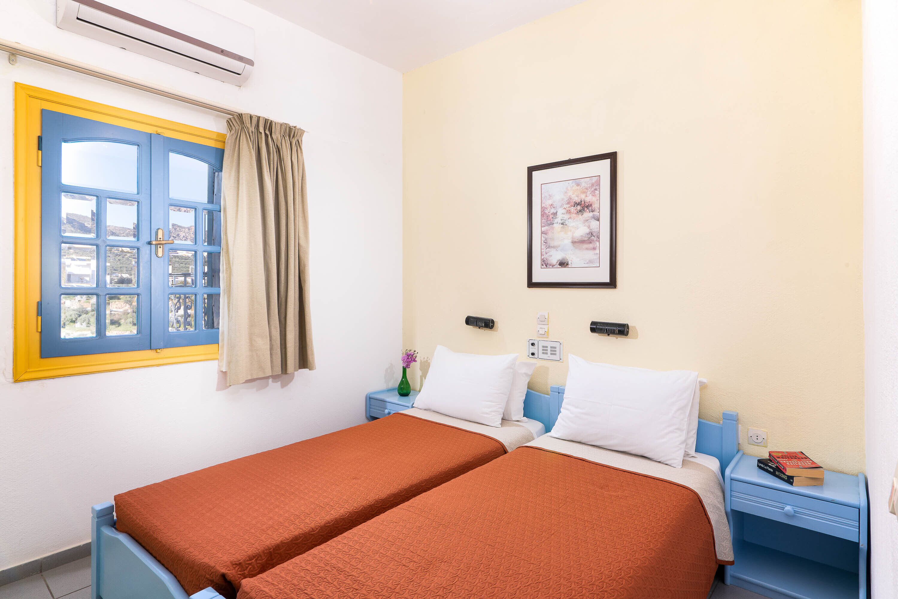 A room with two beds, a window with curtains, a bedside table with a lamp, and a framed picture on the wall.