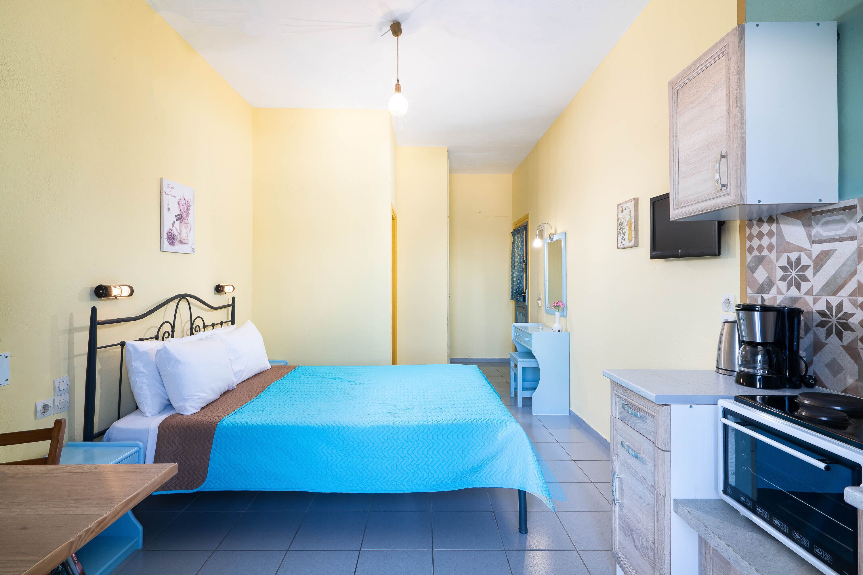 Quarto com cama, mesa de cabeceira e escrivaninha pequena. Possui cozinha compacta com cafeteira e micro-ondas.