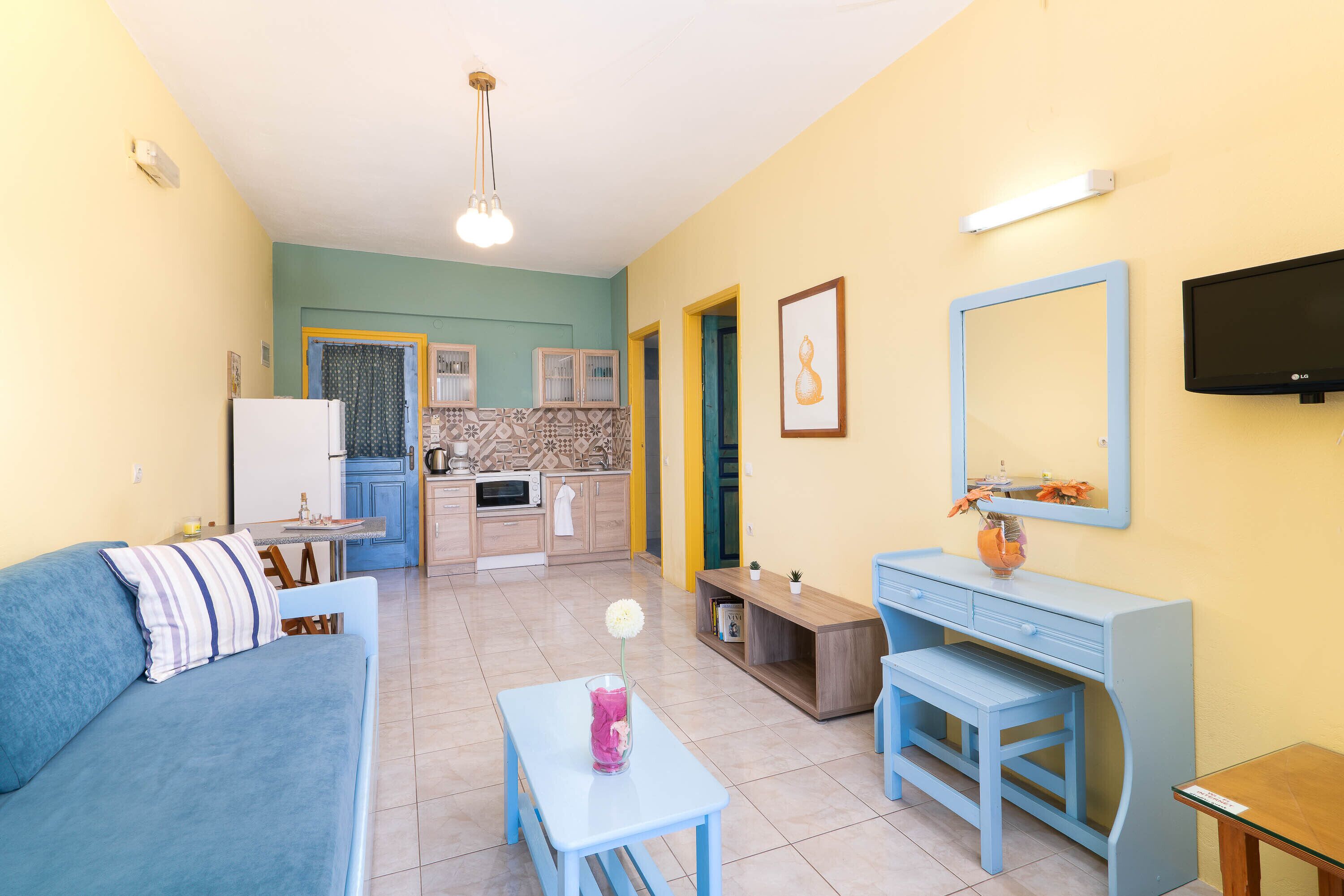 Un espacio habitable compacto con sofá, una mesita con un florero, una kitchenette, un armario de madera y un televisor montado en la pared.