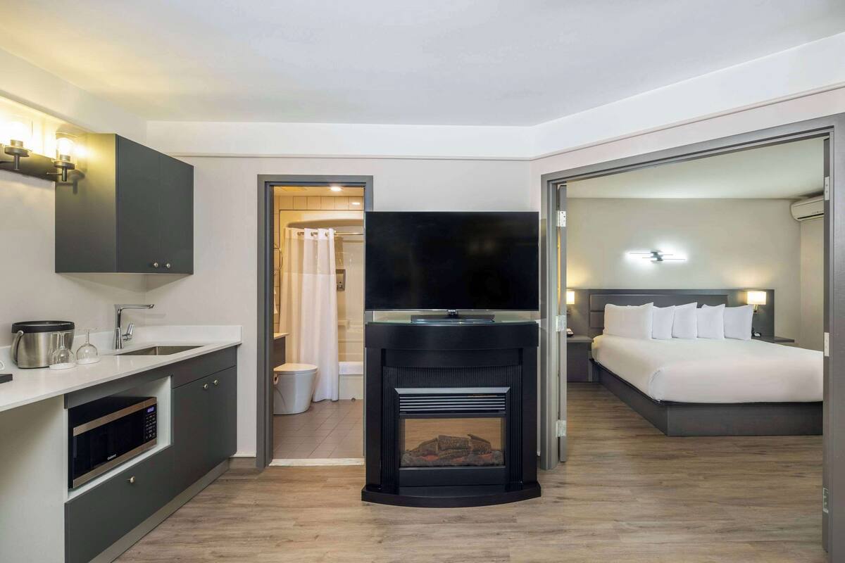 A modern hotel room with a kitchenette, a fireplace, a large bed, and a television.