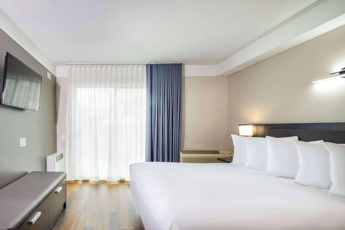 A hotel room with a bed, a desk, a chair, a TV mounted on the wall, and a window with curtains.
