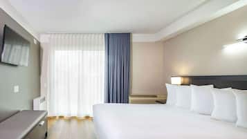 A hotel room with a bed, a desk, a chair, a TV mounted on the wall, and a window with curtains.