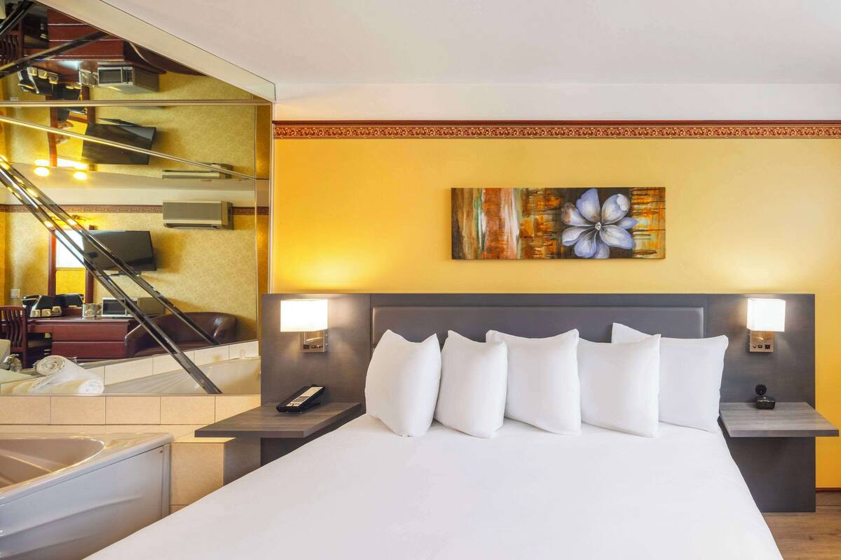 A hotel room with a large bed, two bedside tables, a painting on the wall, and a staircase visible through a glass partition.