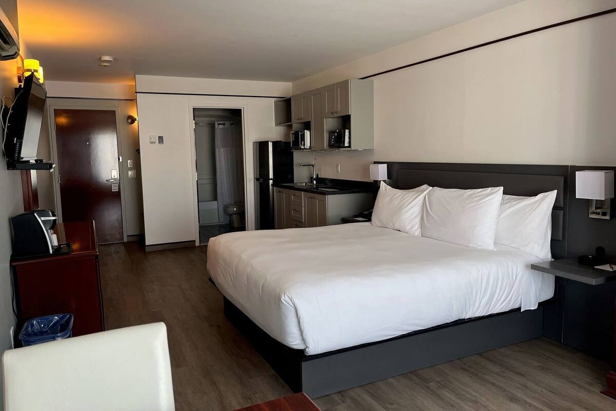 A hotel room with a bed, a desk, a chair, a kitchenette, and a bathroom.