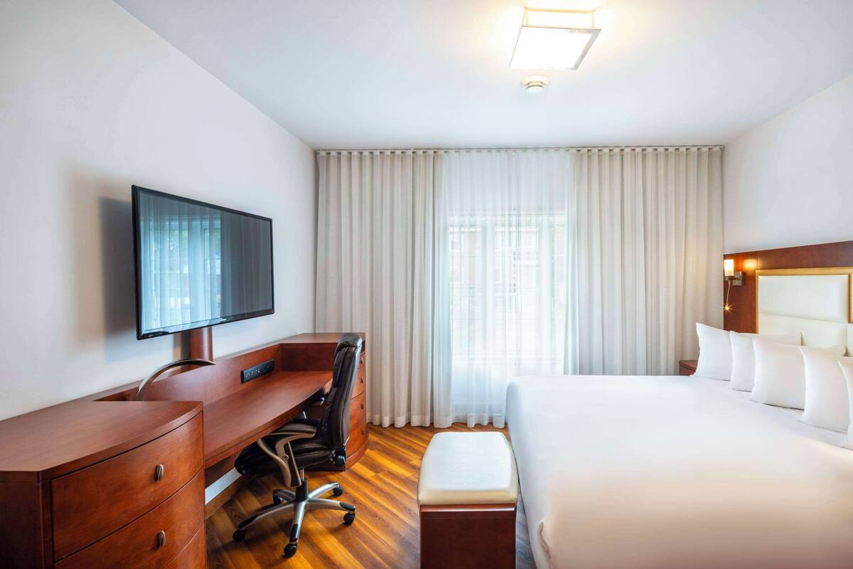 A hotel room with a wooden desk, a television, a bed with white linens, and a large window with curtains.