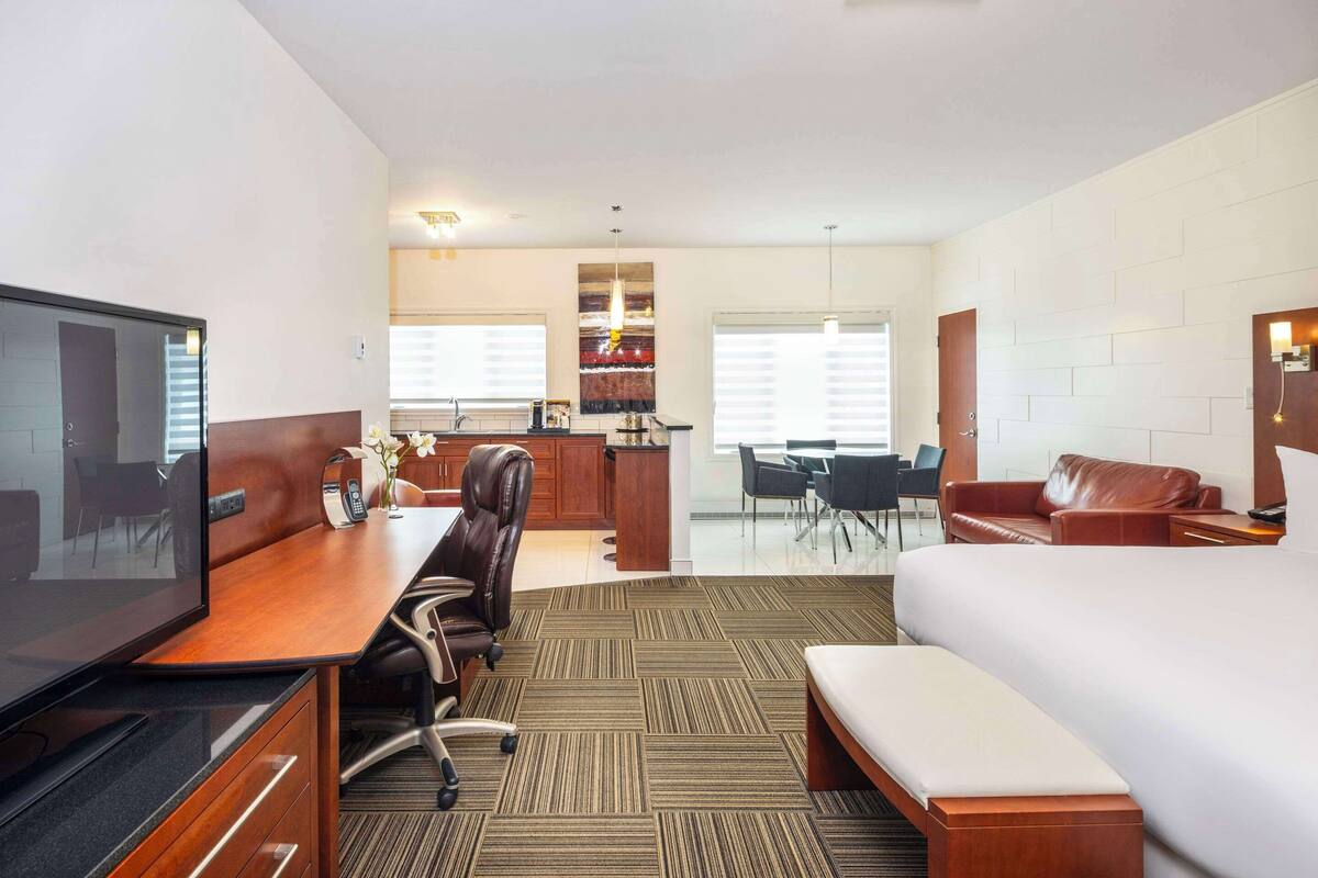 A hotel room with a bed, a desk with a television, a seating area with a sofa and chairs, and a kitchenette in the background.