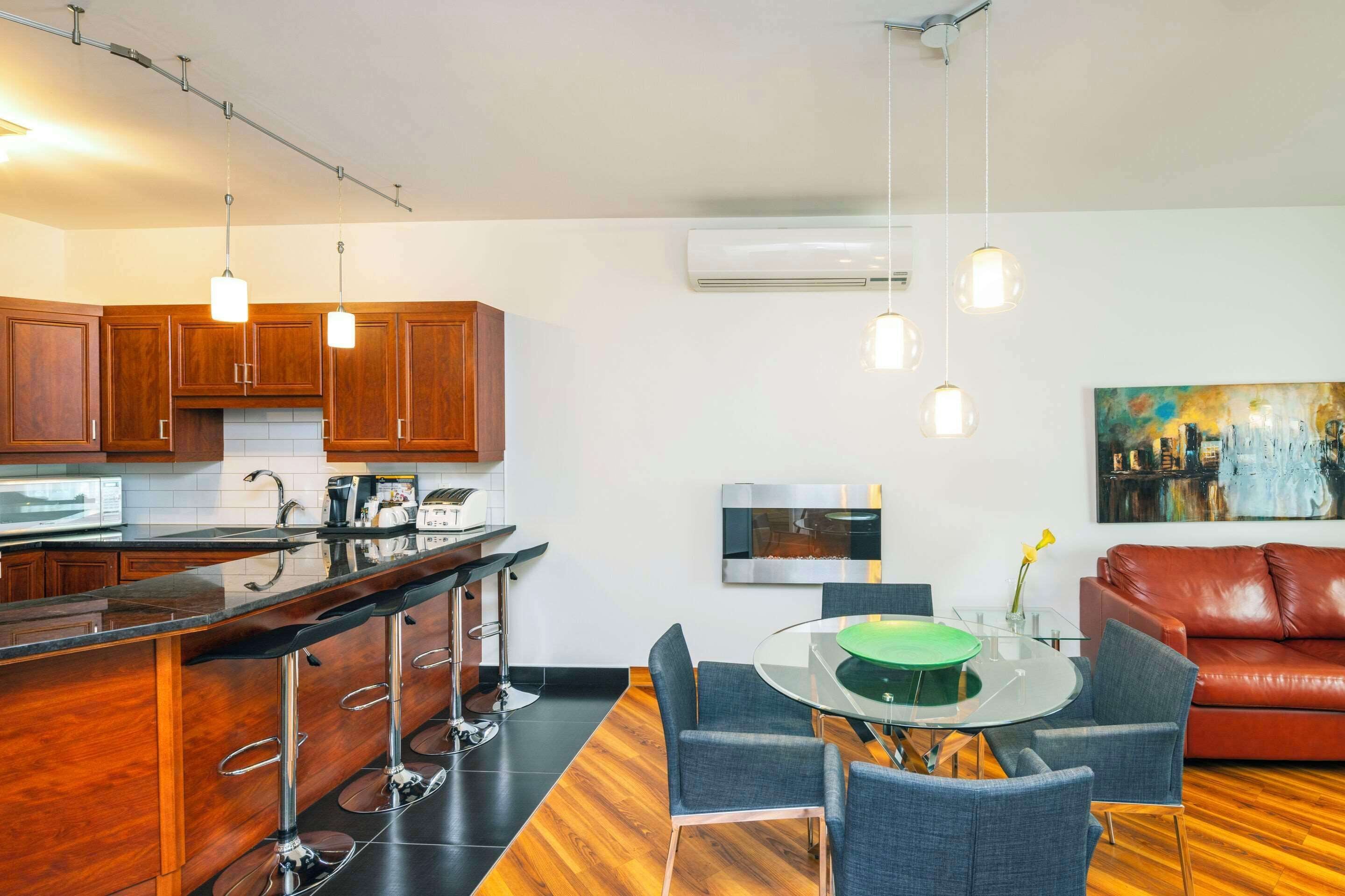 A modern kitchen with wooden cabinets, a black countertop, and a bar area with stools. There is a dining table with chairs and a red leather couch in the living area. An air conditioning unit and a painting are on the wall.