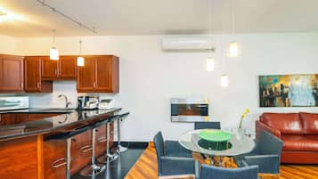 A modern kitchen with wooden cabinets, a black countertop, and a bar area with stools. There is a dining table with chairs and a red leather couch in the living area. An air conditioning unit and a painting are on the wall.