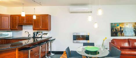 A modern kitchen with wooden cabinets, a black countertop, and a bar area with stools. There is a dining table with chairs and a red leather couch in the living area. An air conditioning unit and a painting are on the wall.