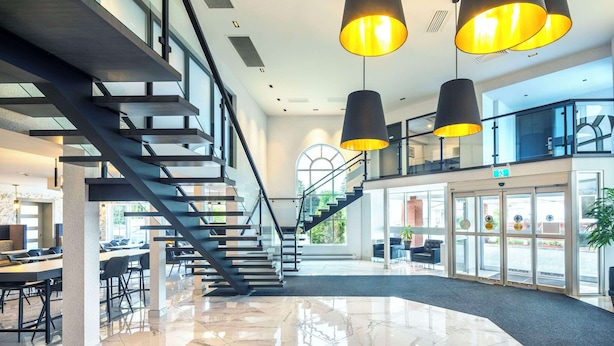 Un hall d’hôtel moderne avec un escalier central, de grandes suspensions et une entrée avec une porte en verre.