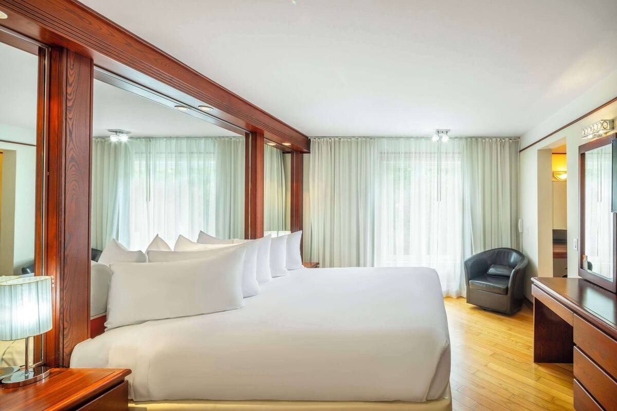 A hotel room with a large bed, a wooden nightstand, a desk, a chair, and a large mirror.
