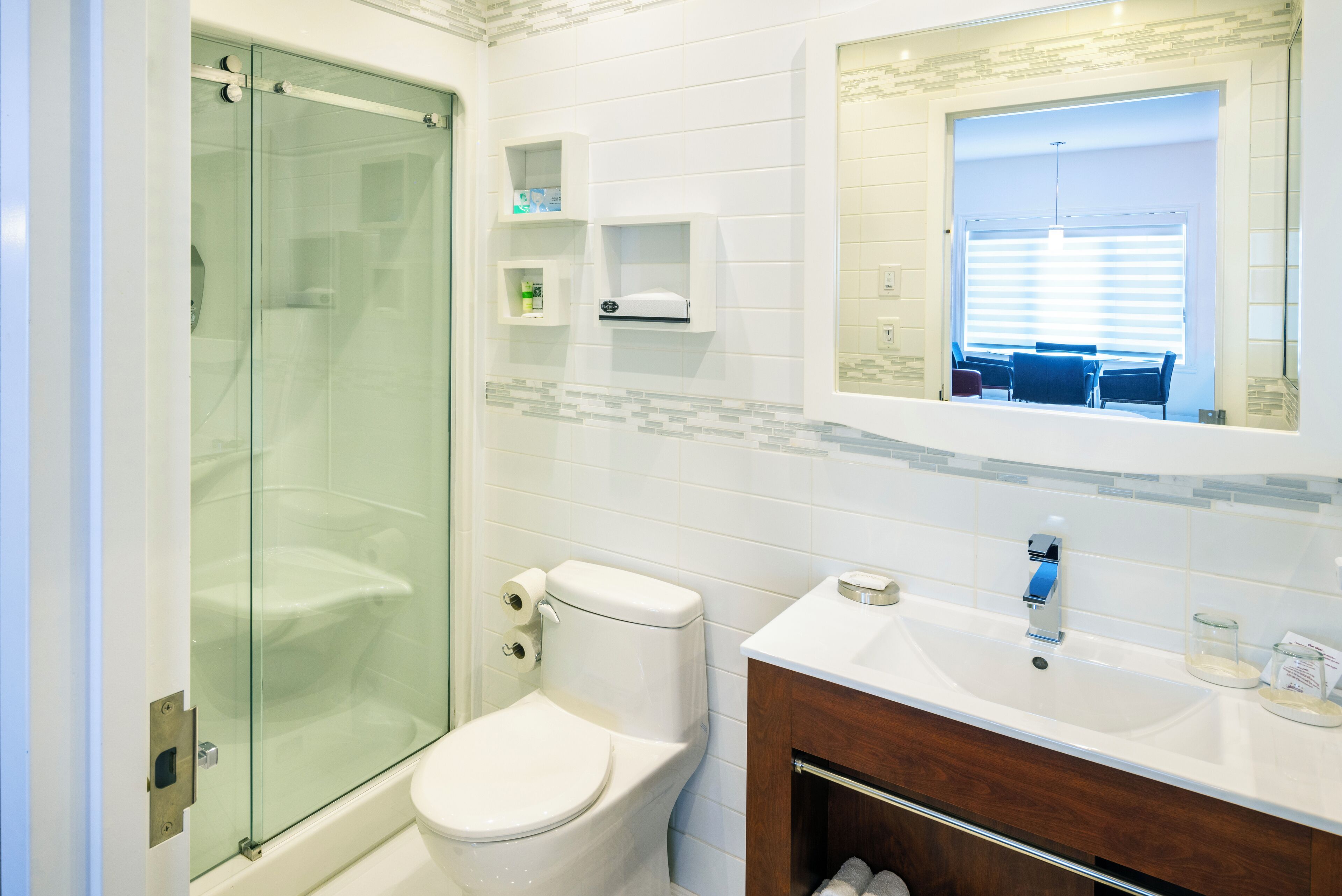 Un baño moderno con cabina de ducha de vidrio, inodoro blanco, lavabo con espejo y un pequeño estante con artículos de aseo.
