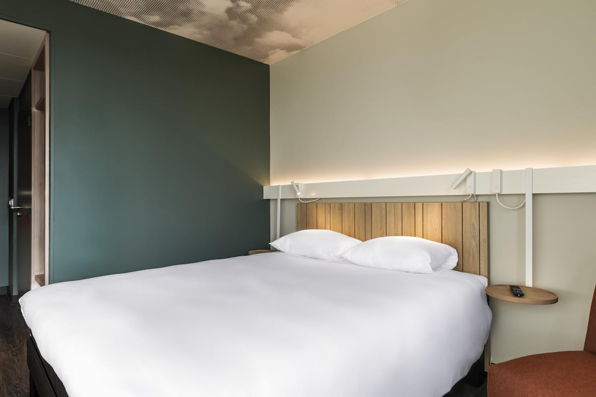 A hotel room with a large bed, a wooden headboard, a bedside table with a remote control, and a brown armchair.
