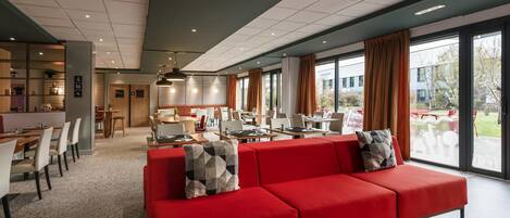 Un moderno vestíbulo de hotel con un sofá rojo, comedor y grandes ventanales que ofrecen vistas al exterior.