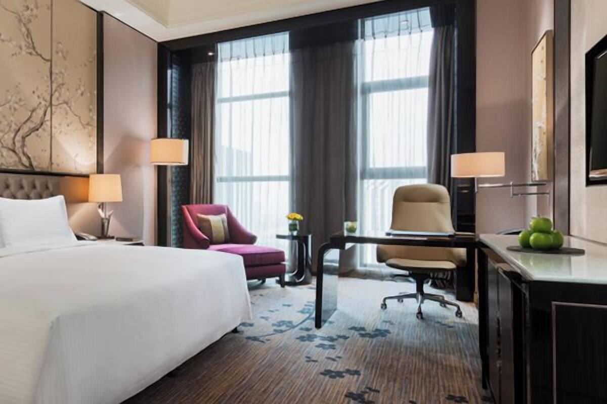 A hotel room with a large bed, a desk, two chairs, and a view of the city through large windows.