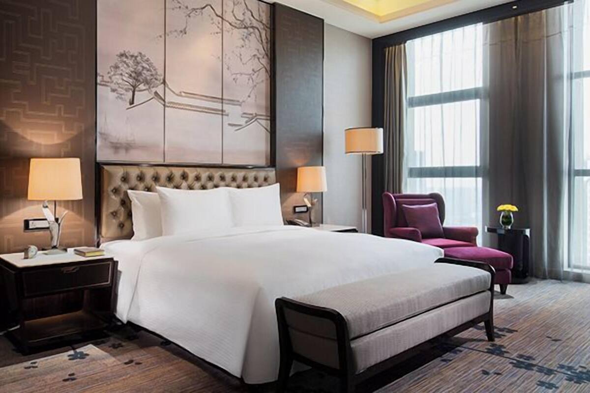 A hotel room with a large bed, two bedside tables with lamps, a chair, and a large window with curtains.