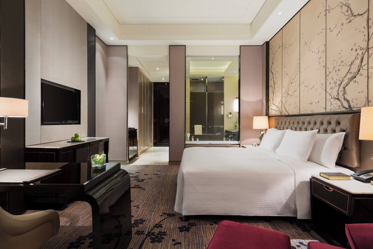 A hotel room with a bed, bedside tables, a desk, a TV, and a large mirror.