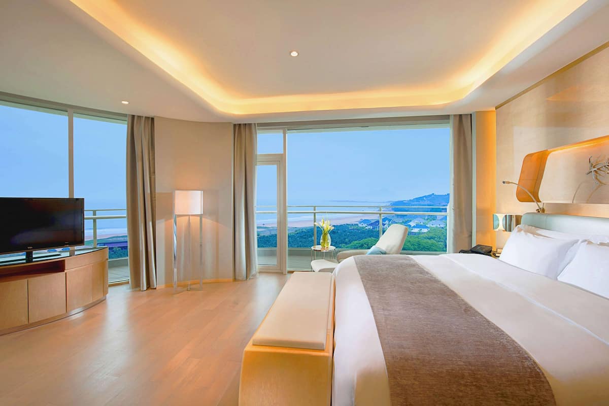 A modern hotel room with a large bed, a television, a desk, and a view of the cityscape.