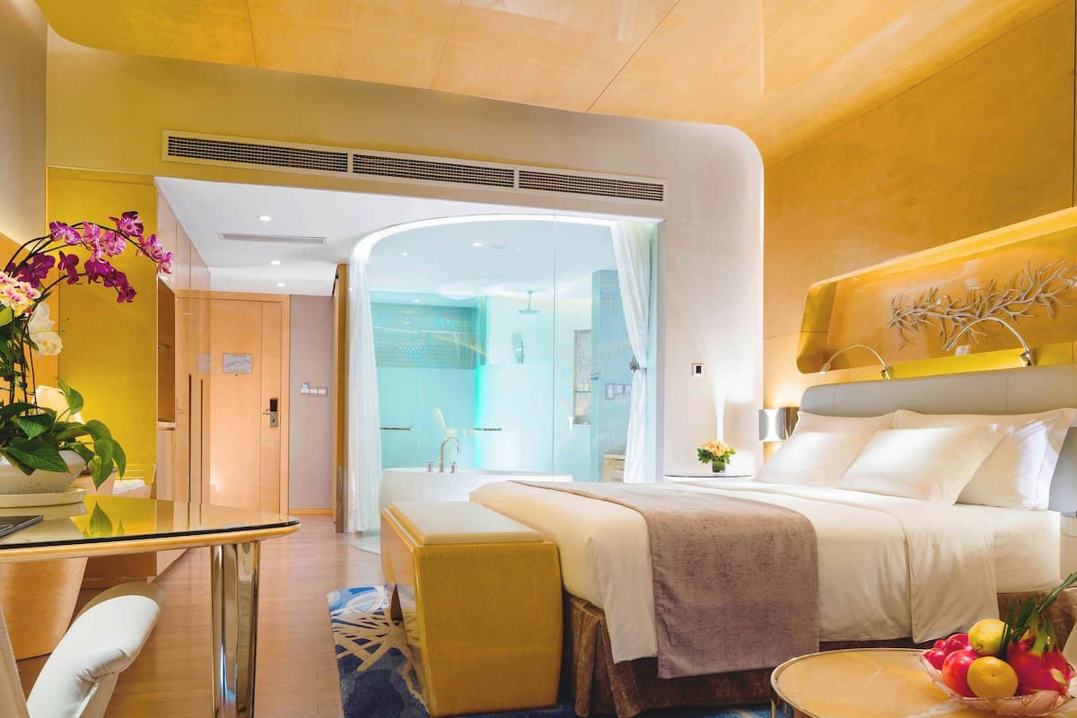 A modern hotel room with a large bed, a glass table, a seating area, and a bathroom visible through an archway.