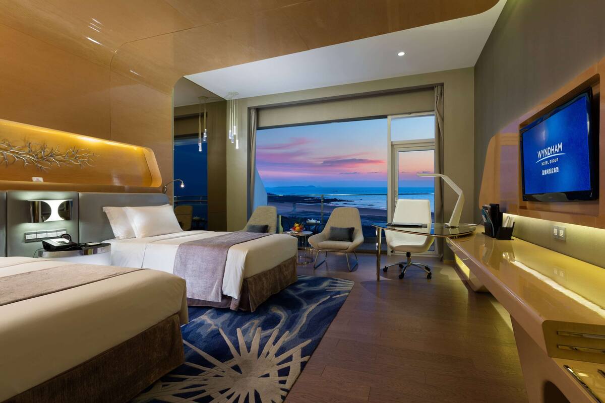 A modern hotel room with a large bed, a desk, a TV, and a view of the sea.