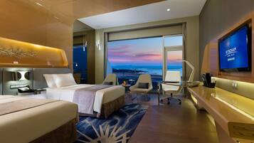 A modern hotel room with a large bed, a desk, a TV, and a view of the sea.