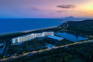 A modern hotel with a large building, a parking area, and a view of the sea and hills. - Wyndham Grand Qingdao (Qingdao)