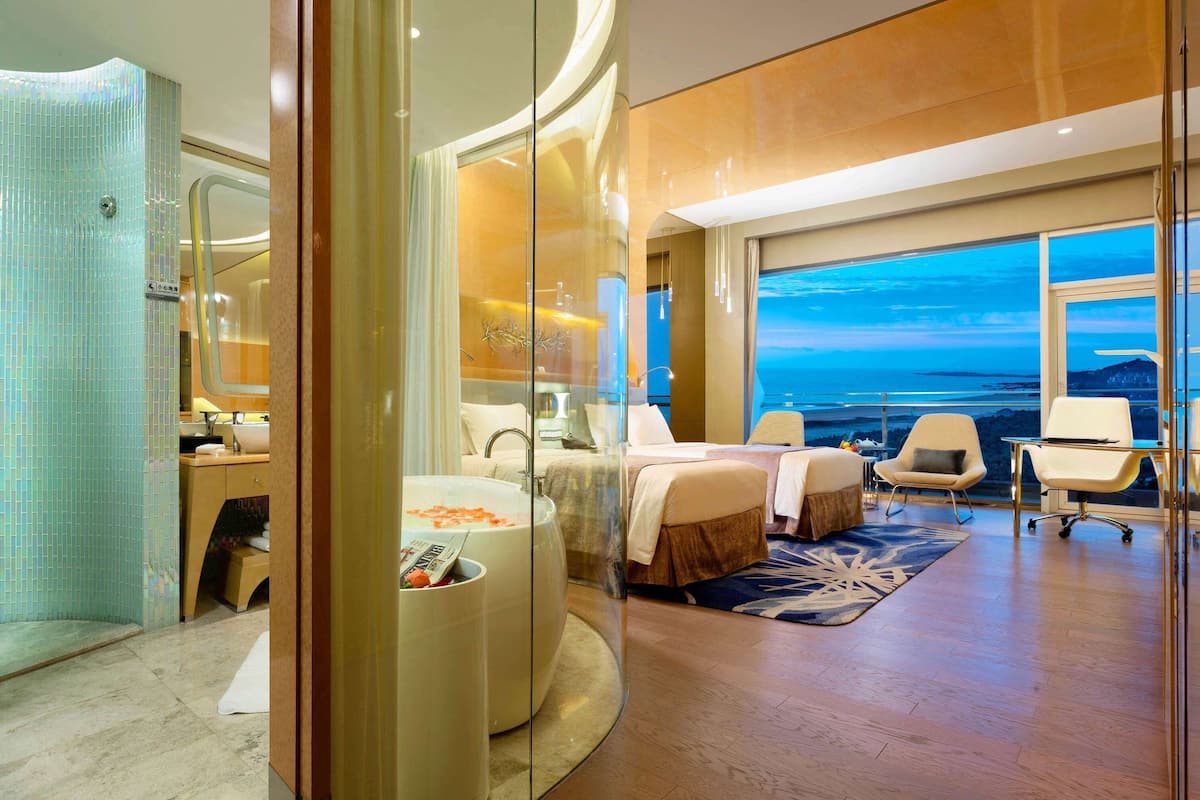A modern hotel room with a large bed, a sitting area with a glass wall, a bathtub, and a view of the ocean.