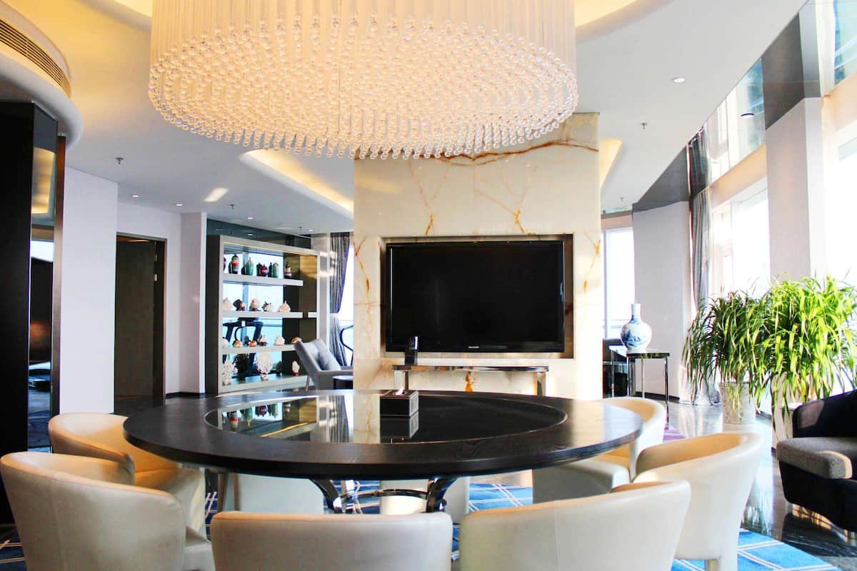 A modern living room with a round black table surrounded by white chairs, a large flat-screen TV mounted on the wall, and a decorative chandelier.