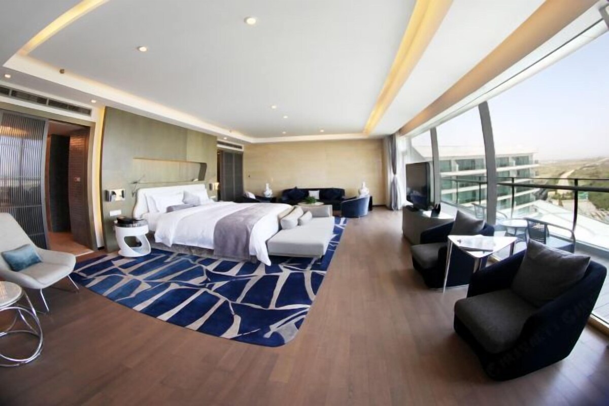 A modern hotel room with a large bed, a sofa, a small table, and a balcony with a view.