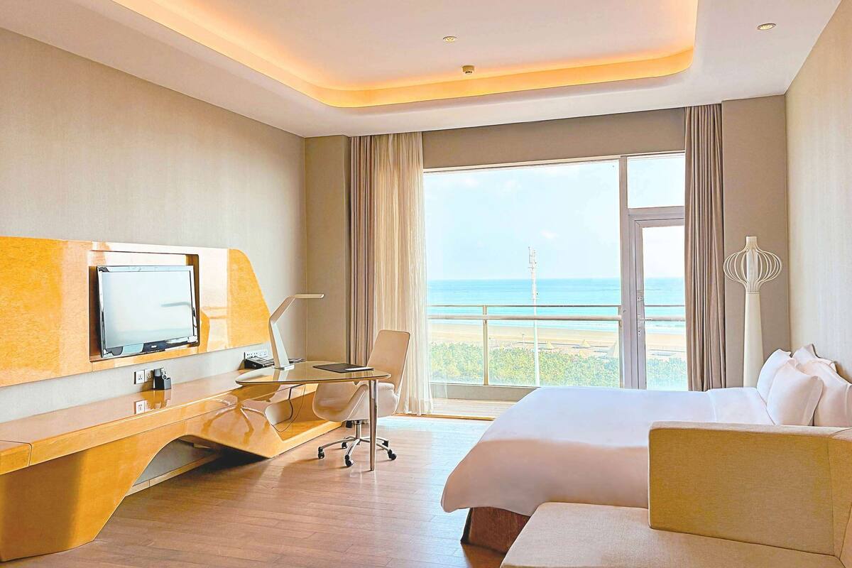 A hotel room with a bed, desk, chair, TV, and a view of the beach.