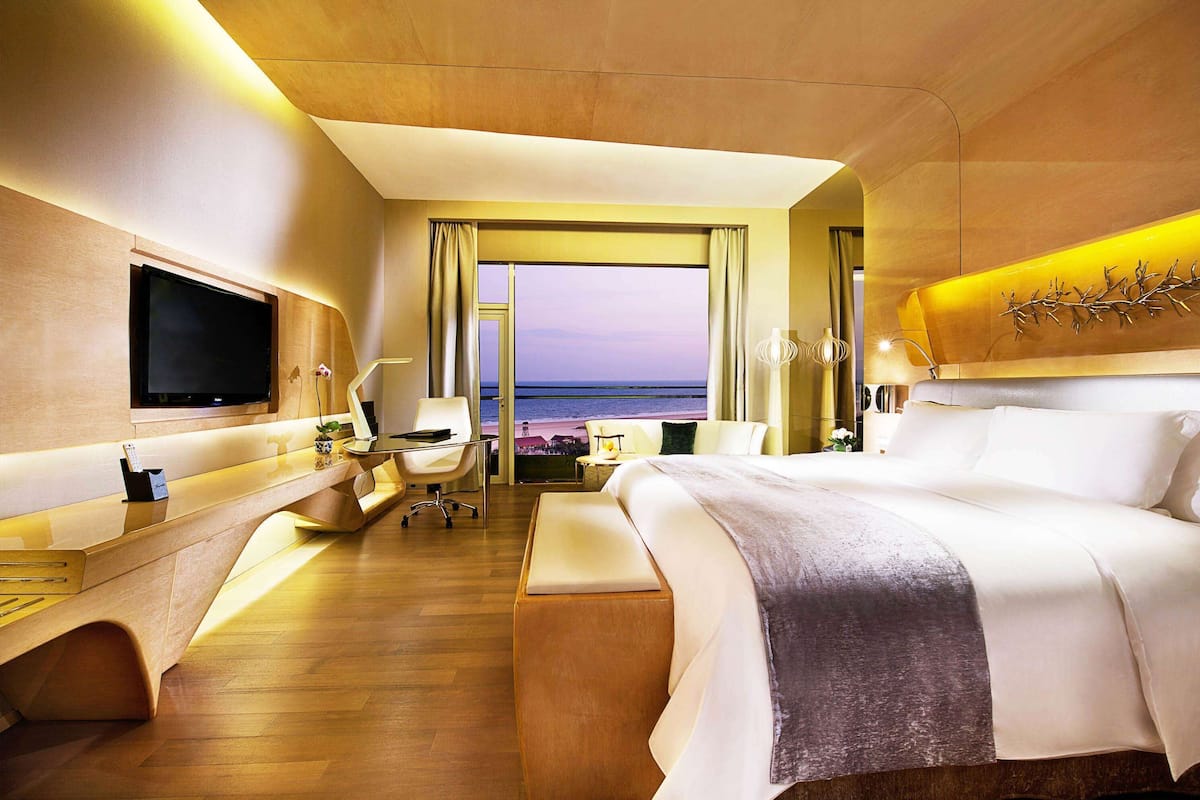 A modern hotel room with a large bed, a desk, a chair, a TV, and a view of the sea.