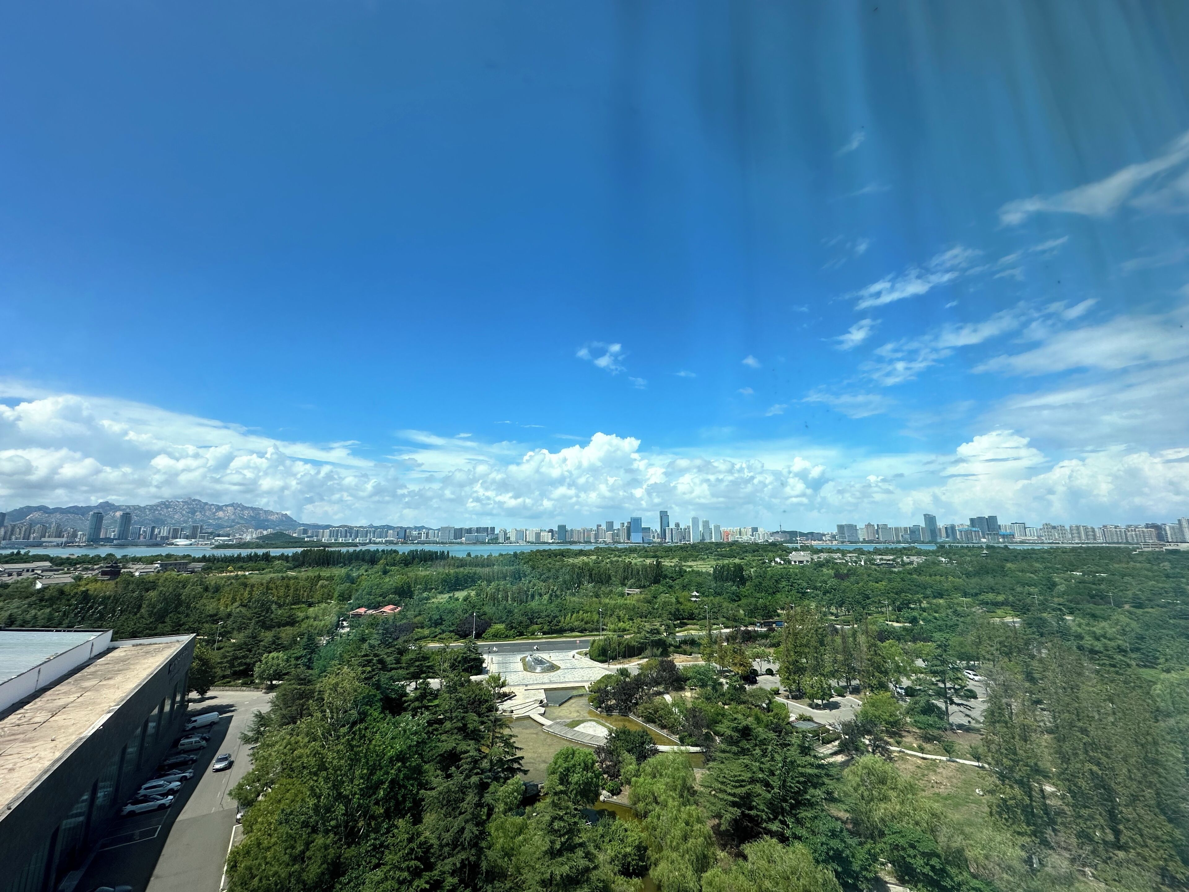 Un paisaje urbano con una mezcla de edificios altos y zonas verdes, un área de estacionamiento y un cielo despejado.