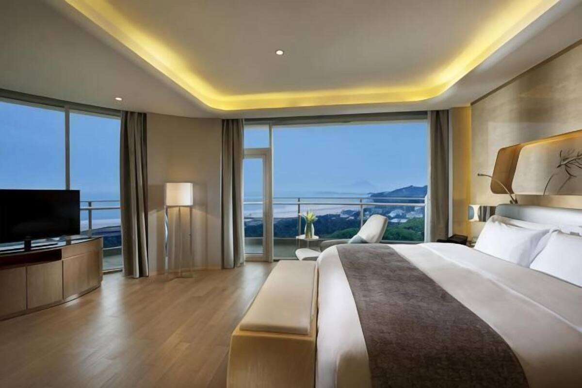 A modern hotel room with a large bed, a flat-screen TV, a bedside table, and a balcony with a view.