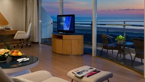 Una habitación de hotel moderna con un televisor grande, asientos cómodos, un comedor y un balcón con vista al océano.