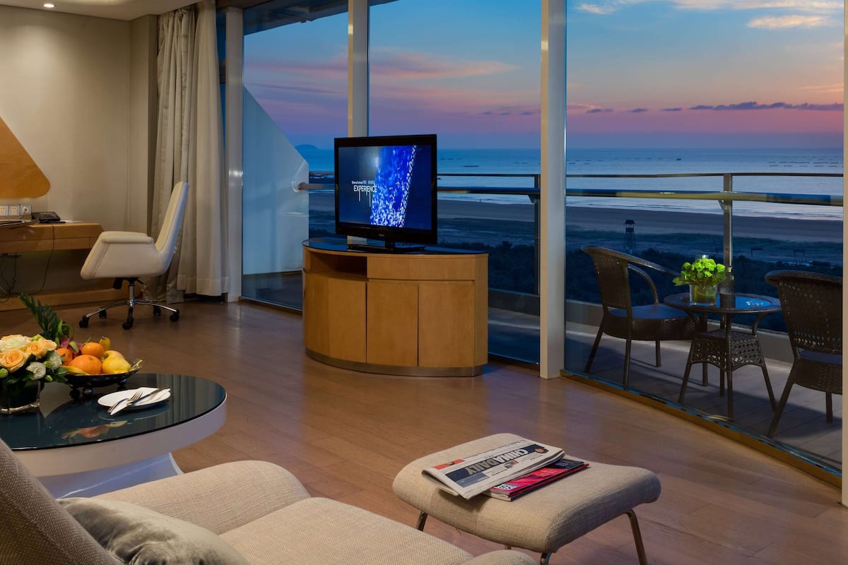A modern hotel room with a large TV, comfortable seating, a dining area, and a balcony with a view of the ocean.