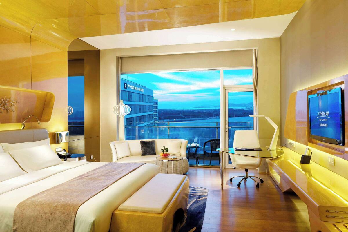 A modern hotel room with a large bed, a sitting area with a glass table, a TV, and a view of the cityscape.