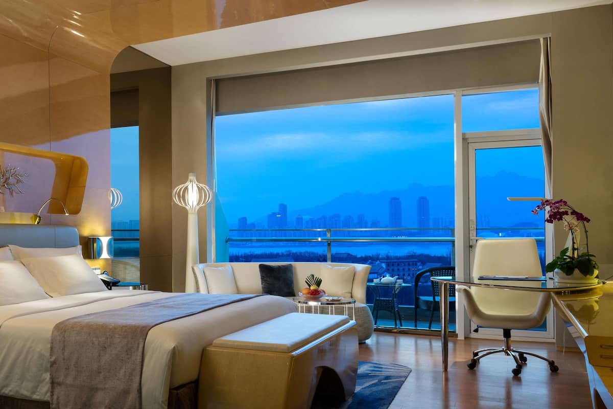 A modern hotel room with a large bed, a desk, a chair, and a balcony with a city view.